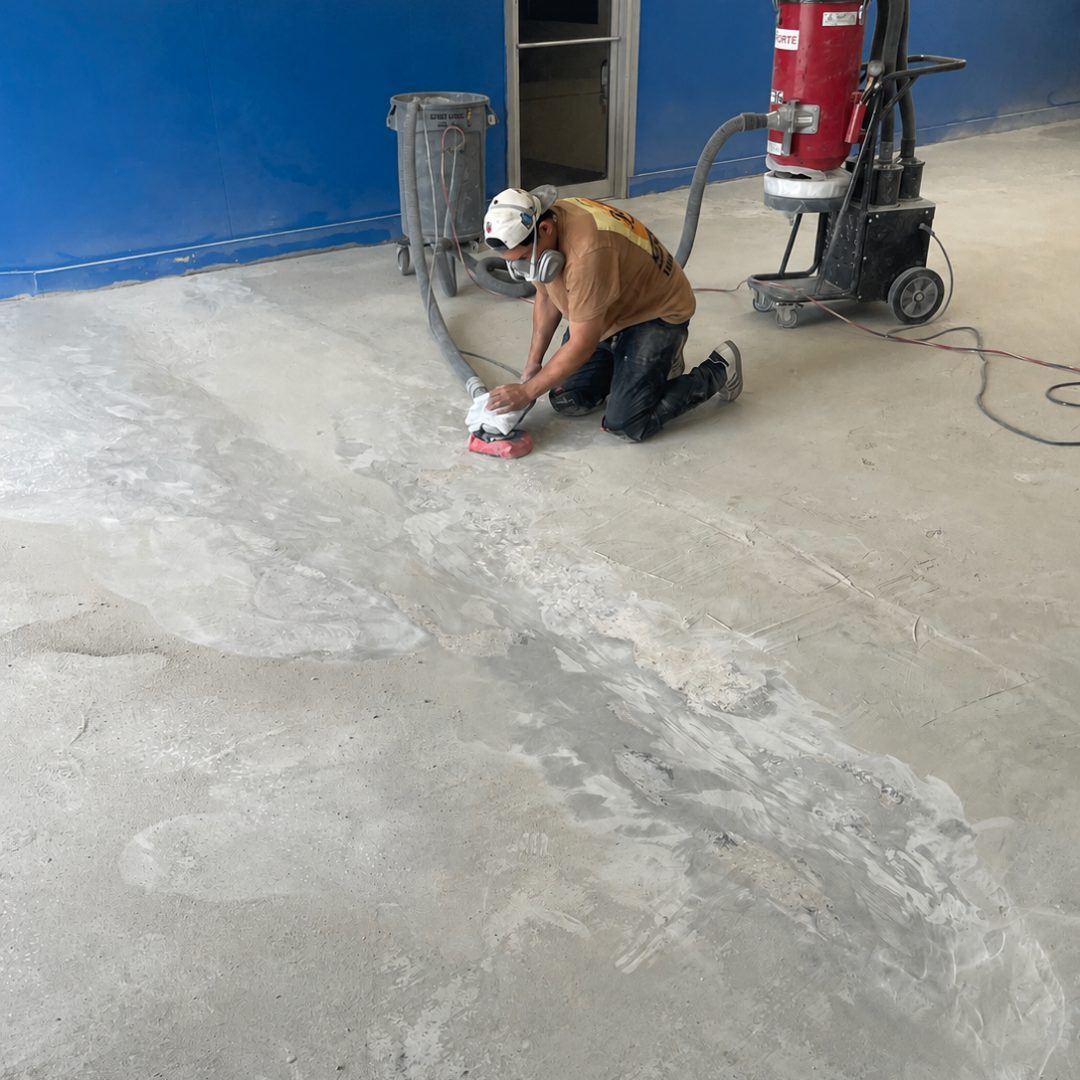 Crew member in a respirator hand-grinding the edges of a concrete floor with a HEPA dust extractor running alongside
